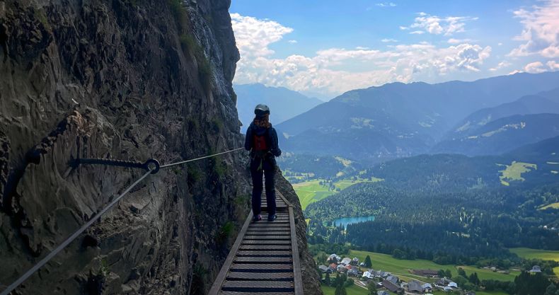 Eine Bergsteigerin in Kletterausrüstung steht auf einem schmalen Klettersteig an einer Felswand links im Bild, die nach rechts unmittelbar neben der Bergsteigerin vertikal abfällt. Im Vordergrund ist die Sicherungsvorrichtung in der Felswand zu sehen. Die